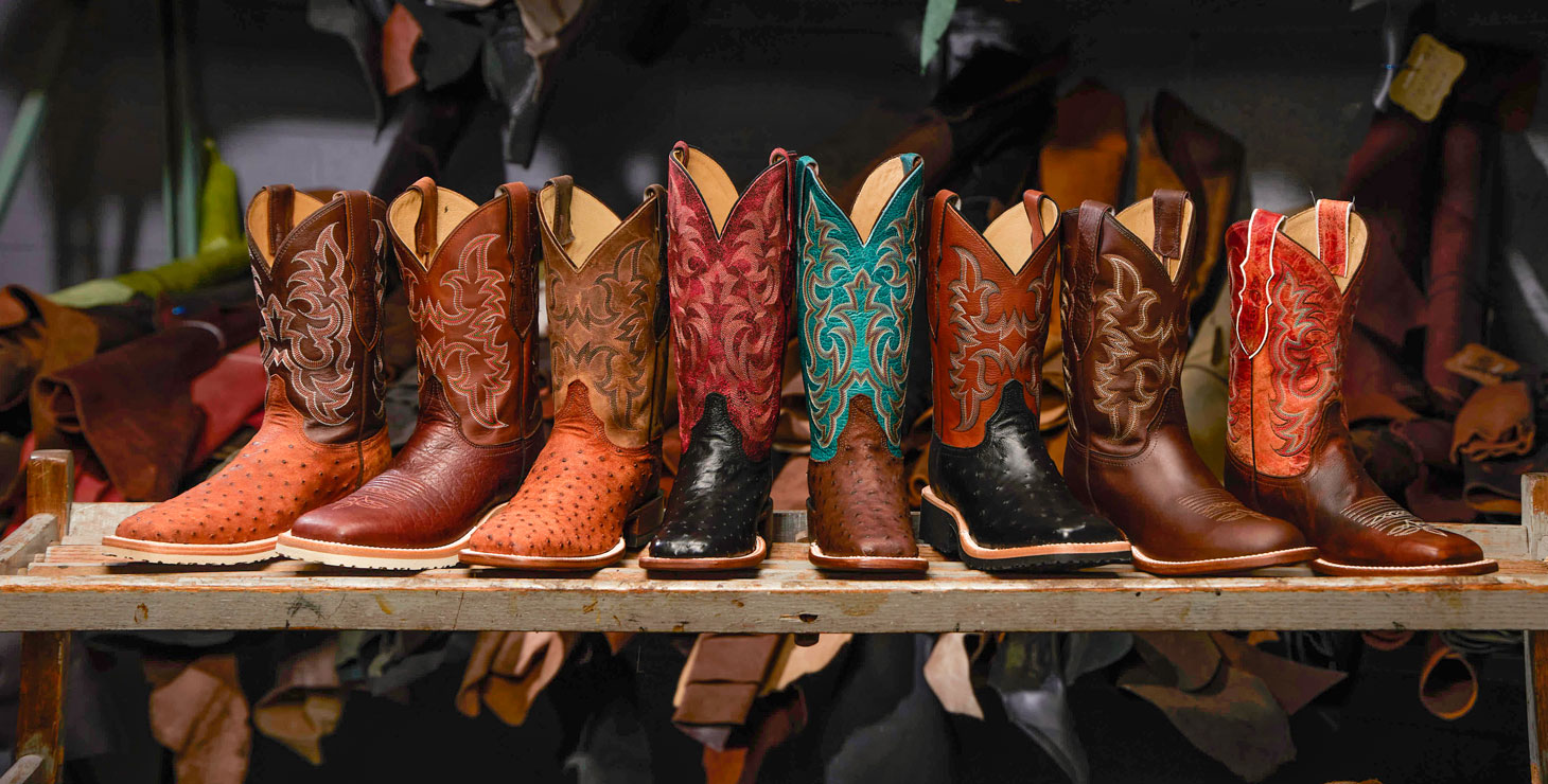 A photo of men’s and women’s cowboy boots lined up in a row with different colors, skins, and styles of brown, tan, maroon, turquoise, burnt orange, and red shafts and brown, tan, and black counters, all posed in front of shelves of rolled up leather skins. 